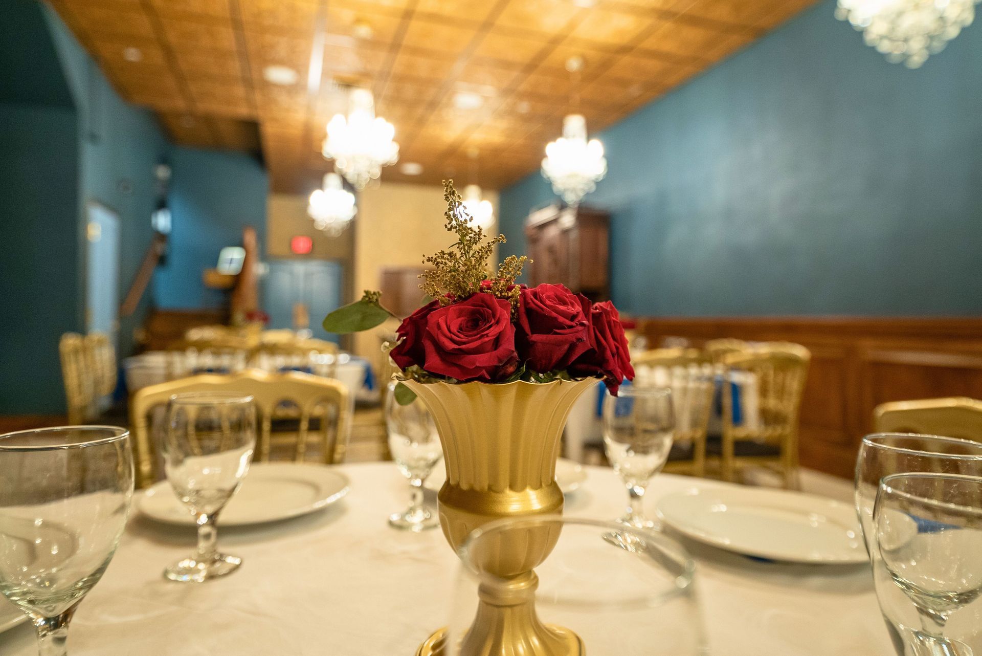 Red roses in a gold vase on a set dining table in a room with blue walls, wooden ceiling, and hanging crystal lights.