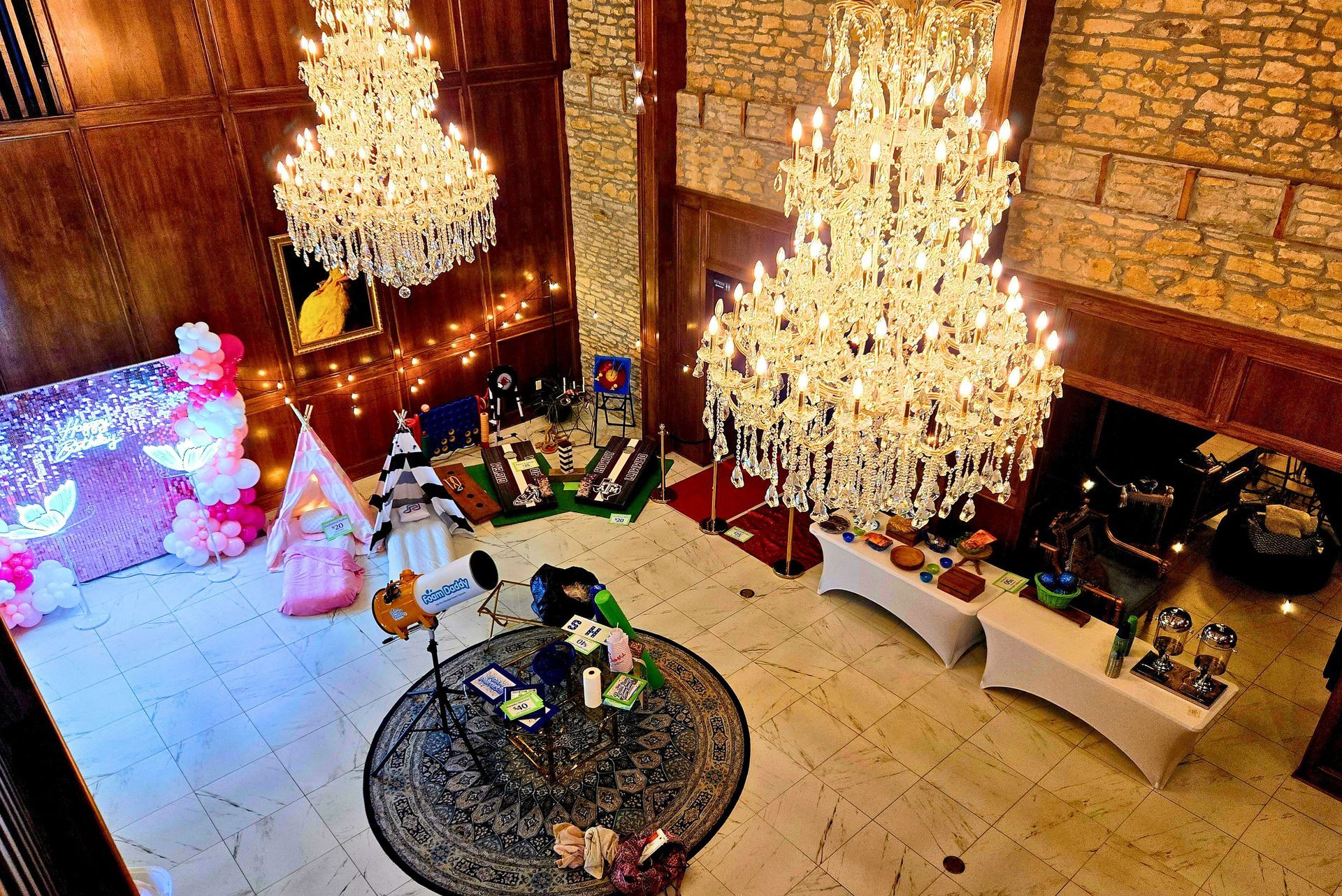 An overhead view of an ornate, high-ceilinged room decorated for a party with chandeliers, pink balloons, and teepees.