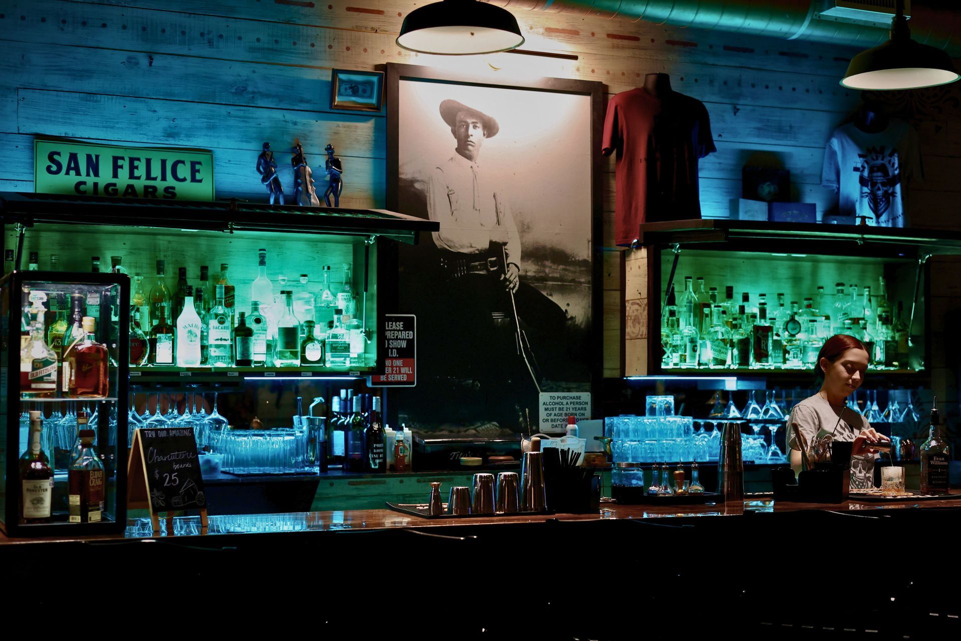 A dimly lit bar with blue/green lighting, featuring a large framed photo of Frank Hamer, bottles on display, and wooden counters.