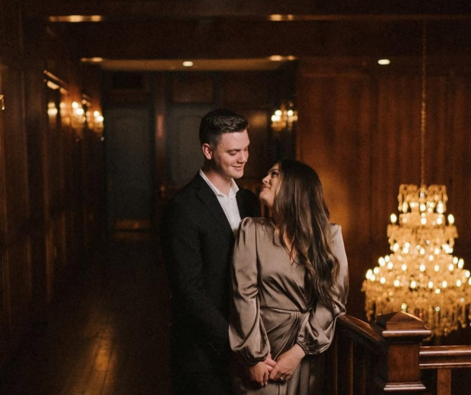 A couple looks at each other while standing on a staircase in a wood-paneled room illuminated by a large chandelier.