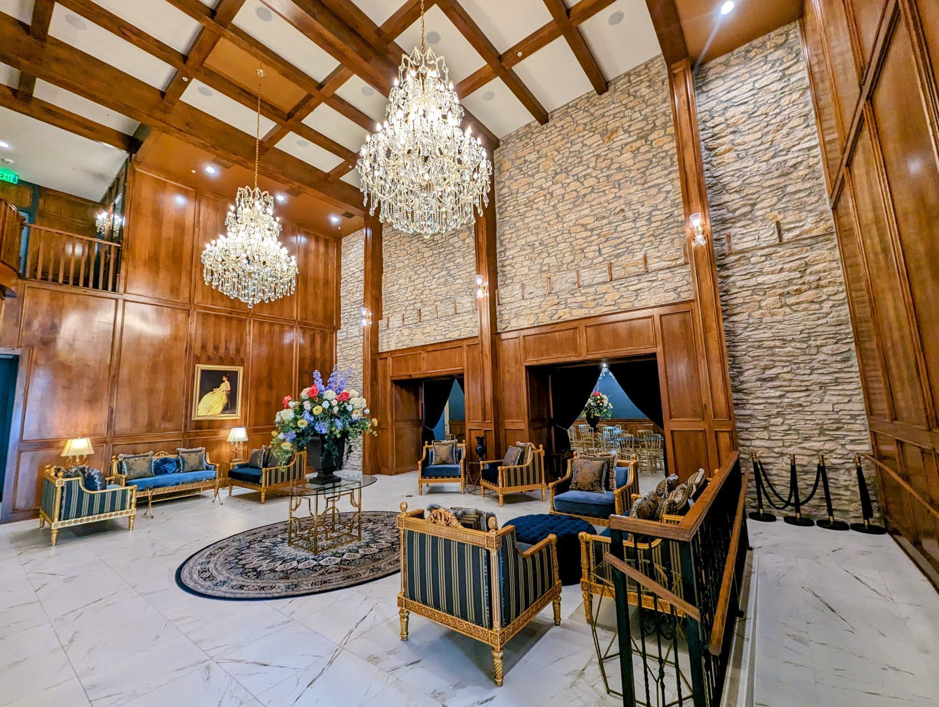 A grand, high-ceilinged lobby with paneled wood walls, stone accents, marble floors, two chandeliers, and plush furniture.
