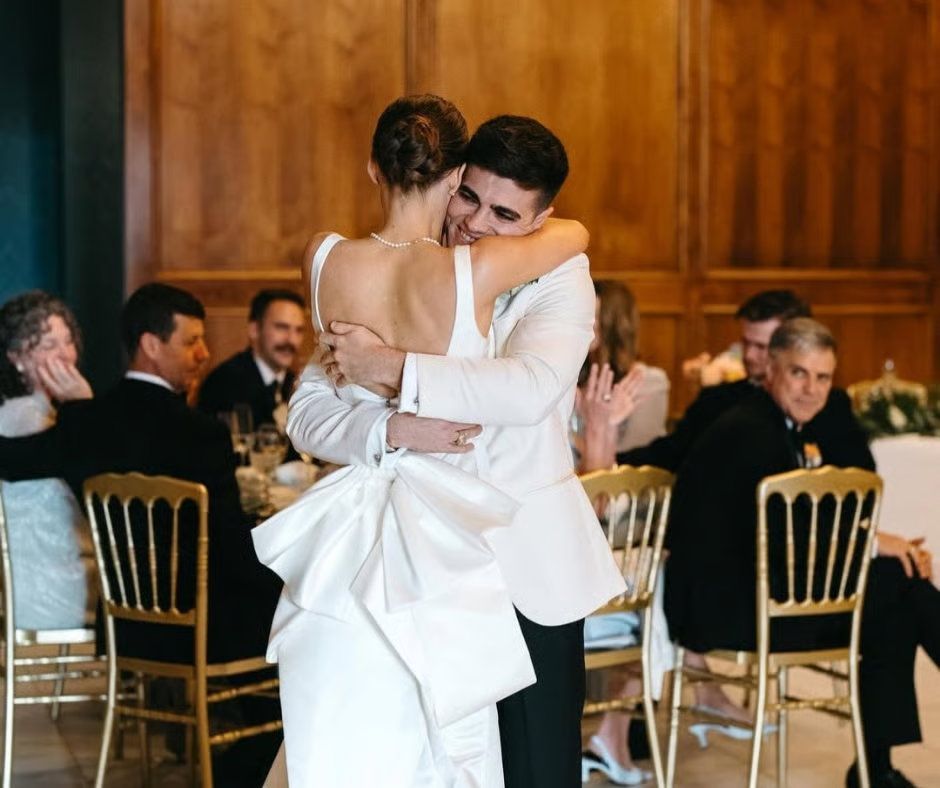 A couple embraces during their wedding reception in a wood-paneled room as guests sit at tables in the background.
