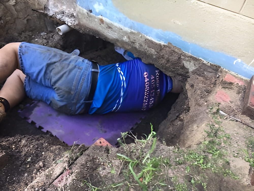 A Man in a Blue Shirt is Laying in a Hole in the Ground — De Rose Plumbing In Bayview Heights, QLD