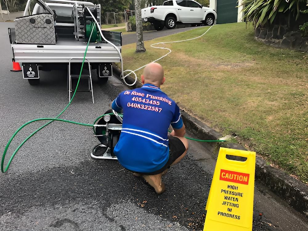 A Man is Kneeling on the Side of the Road Next to a Yellow Caution Sign — De Rose Plumbing In Bayview Heights, QLD