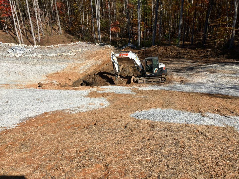Yellow excavator on a flatbed truck parked on asphalt with traffic cones, blue sky, and industrial buildings in background.