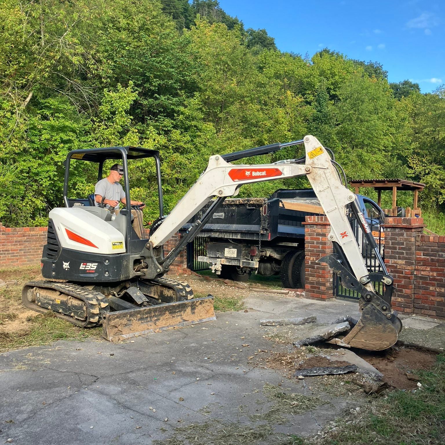 Two excavators, one yellow and one black, on a dirt mound, with trees and a blue sky in the background.