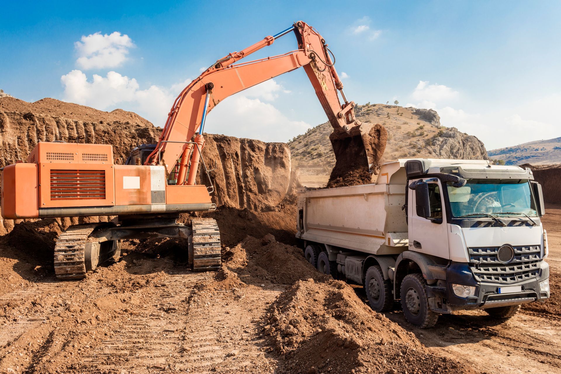An orange excavator loading a white dump truck with dirt at a construction site under a blue sky.
