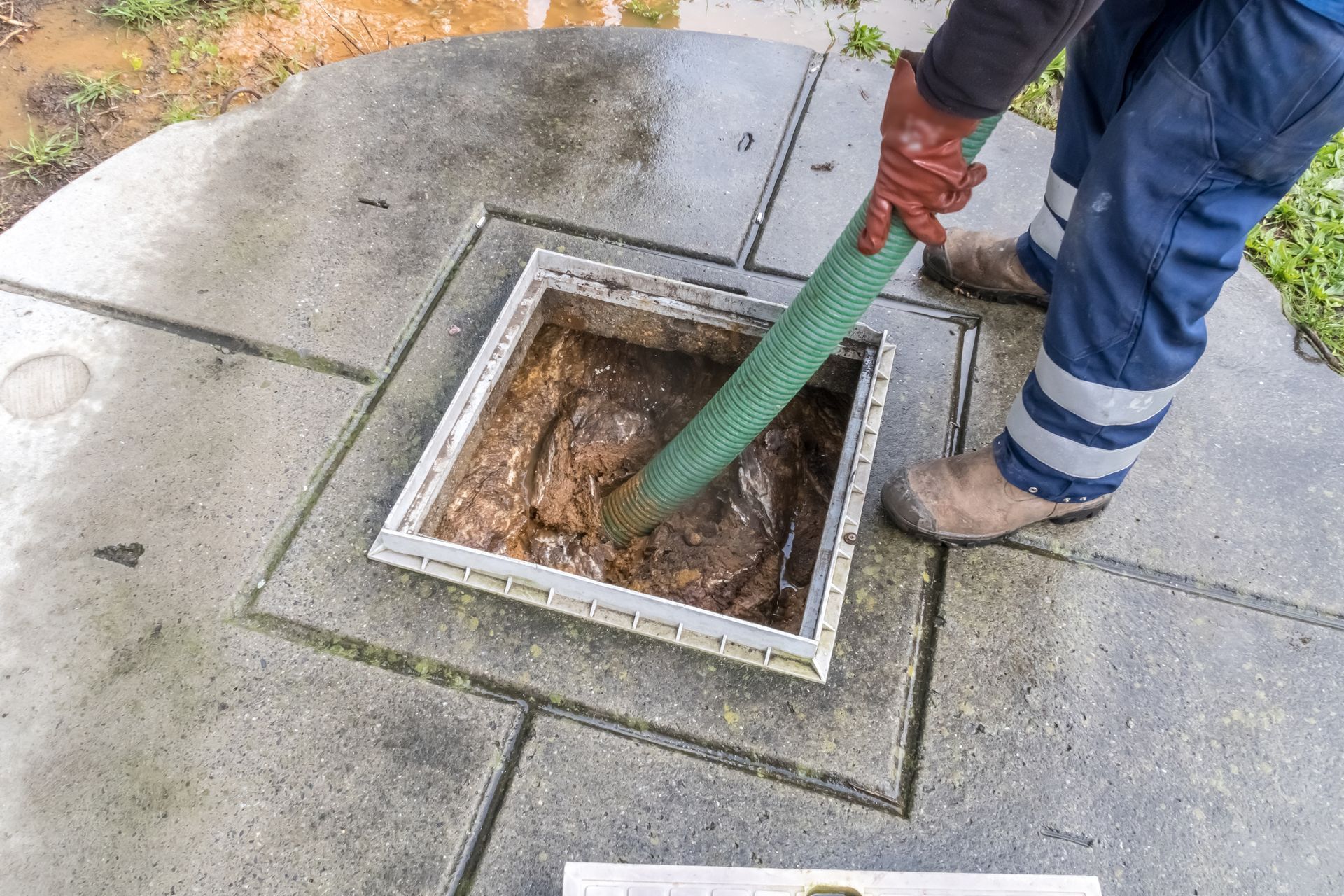 Person in work clothes using a hose to clean a septic tank access, surrounded by concrete.
