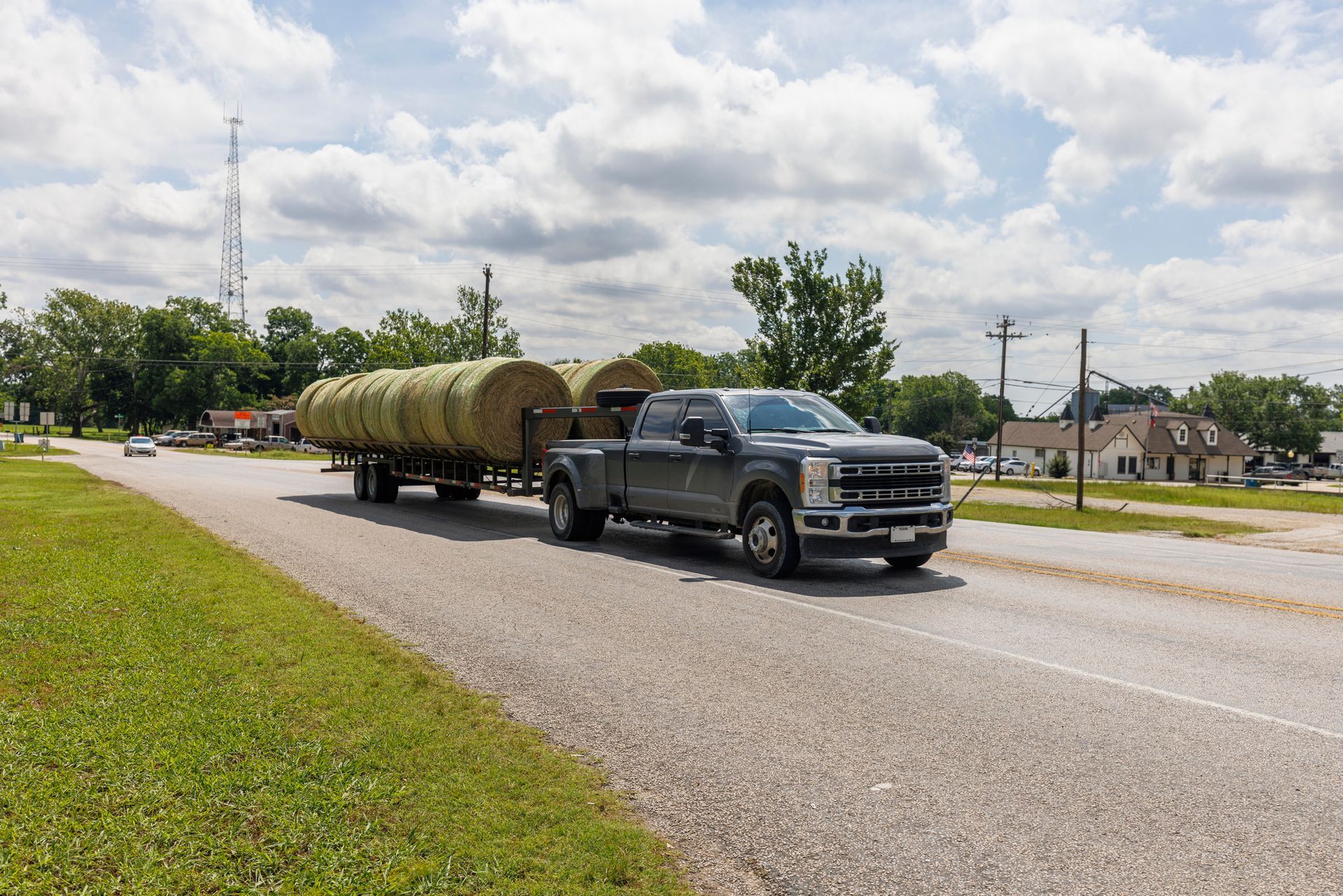 Truck hauling two hay bales on a trailer on a road, with trees and houses in the background.