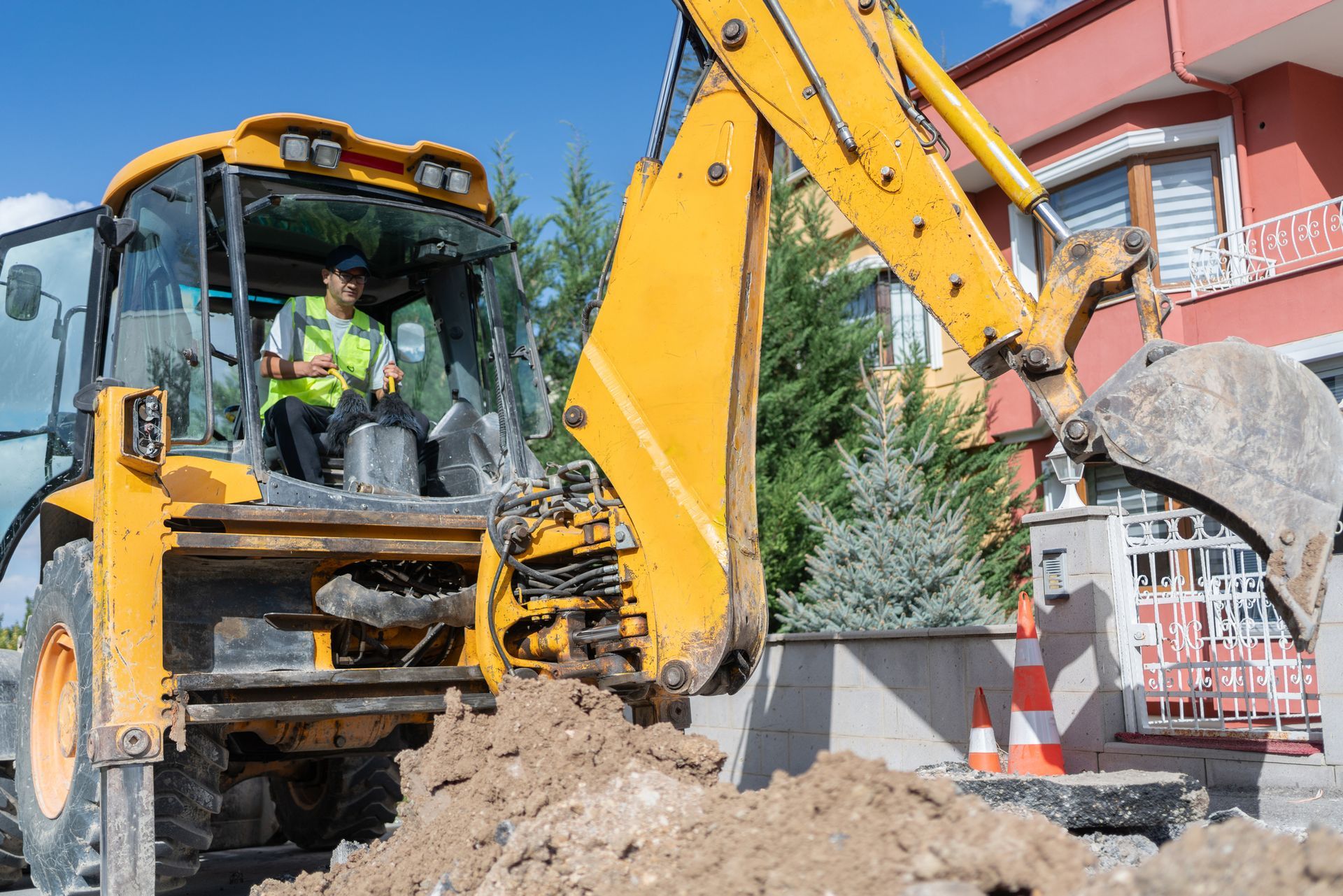 A yellow backhoe digging in front of a red building, with a person operating the machinery.