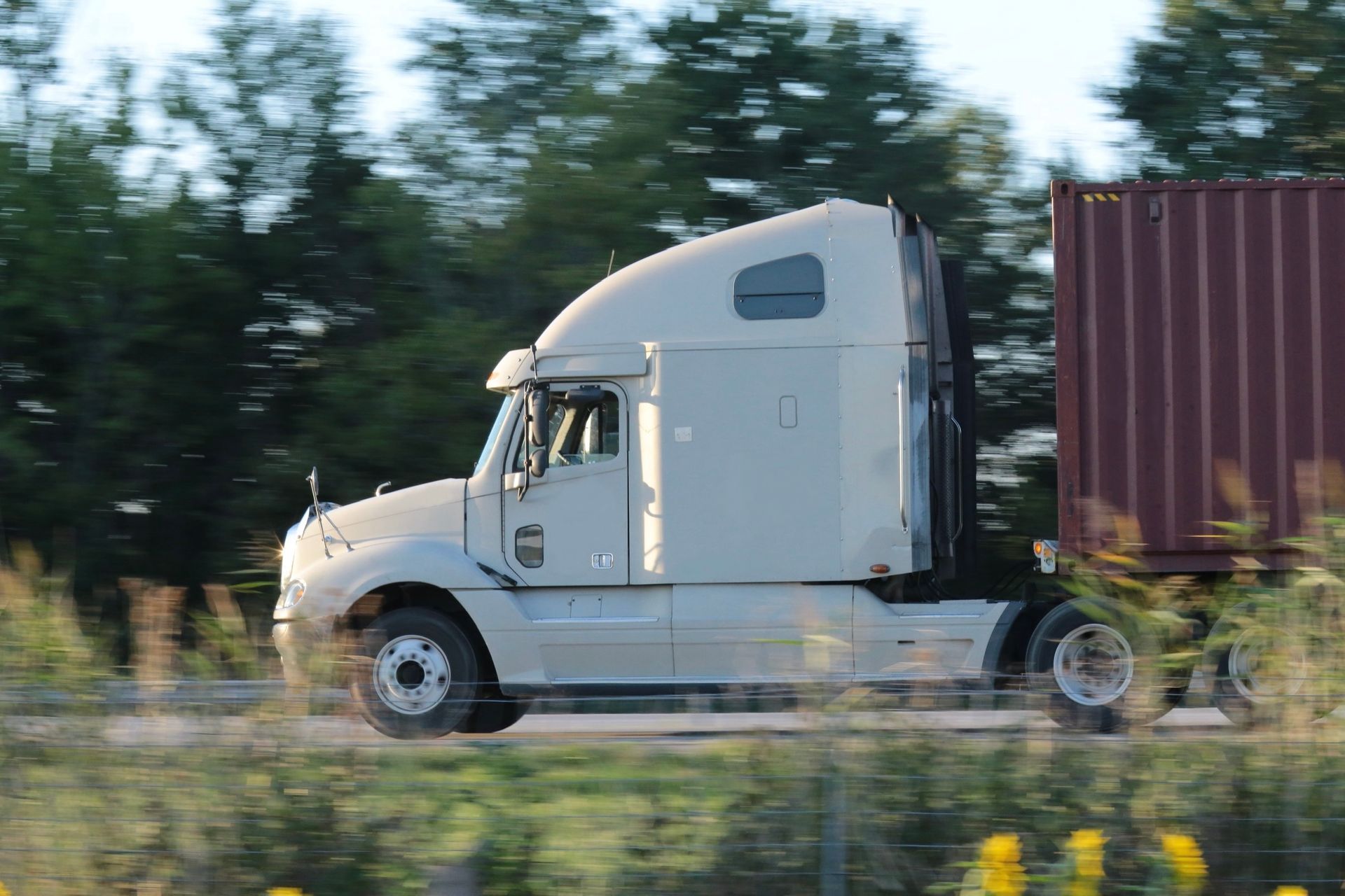 White semi-truck with a maroon cargo container traveling on a highway, green trees in the background.