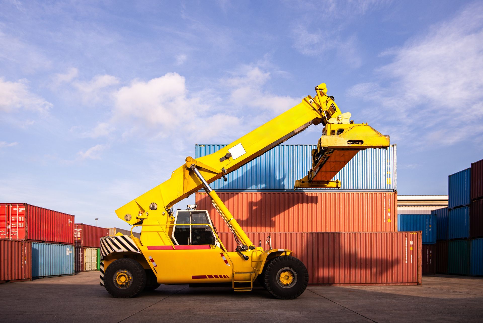 Yellow forklift lifting a shipping container at a port, against a blue sky with clouds.