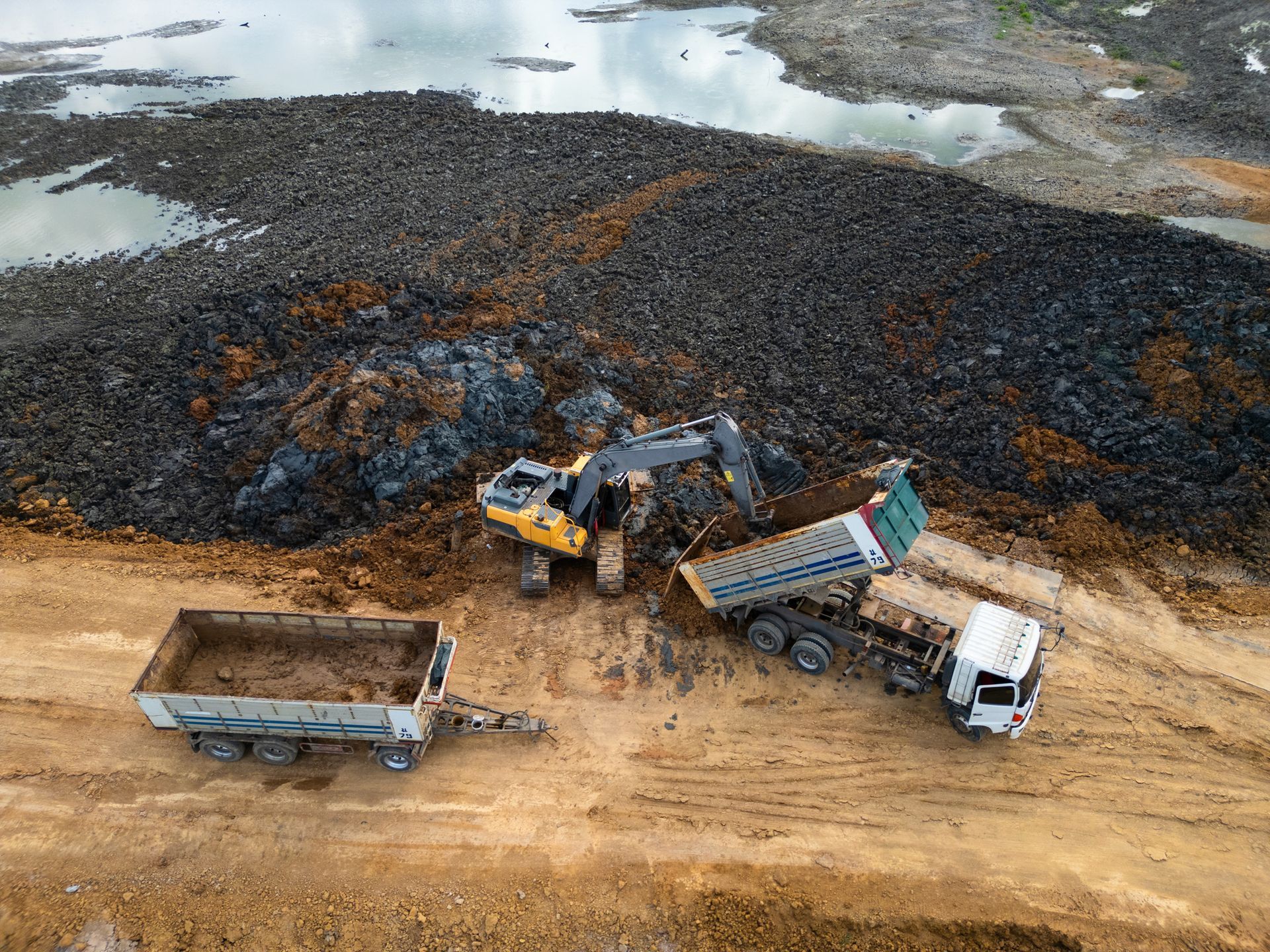 Excavator loading dirt into dump trucks at a construction site. Brown earth, blue sky, and water visible.