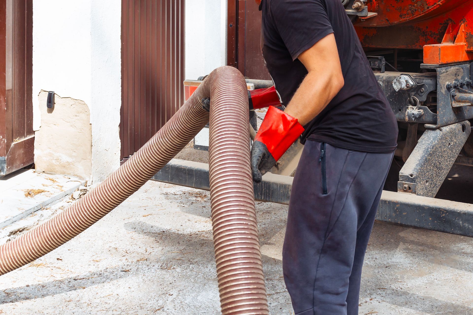 Man in red gloves holding a large hose near a building, next to a truck.
