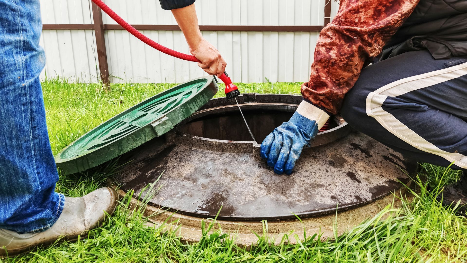 People cleaning a septic tank. One holds a hose; the other, wearing gloves, works inside. Outdoors, green grass.