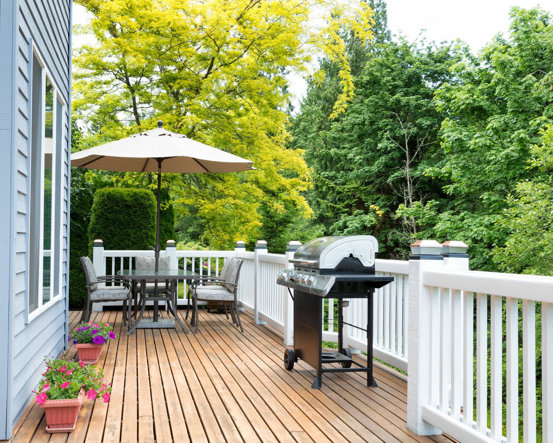 Terrasse en bois avec barbecue, table, chaises et parasol ; entourée d'une rambarde et d'arbres.