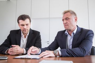 Two businesspeople in suits reviewing documents at a conference table, one pointing at papers