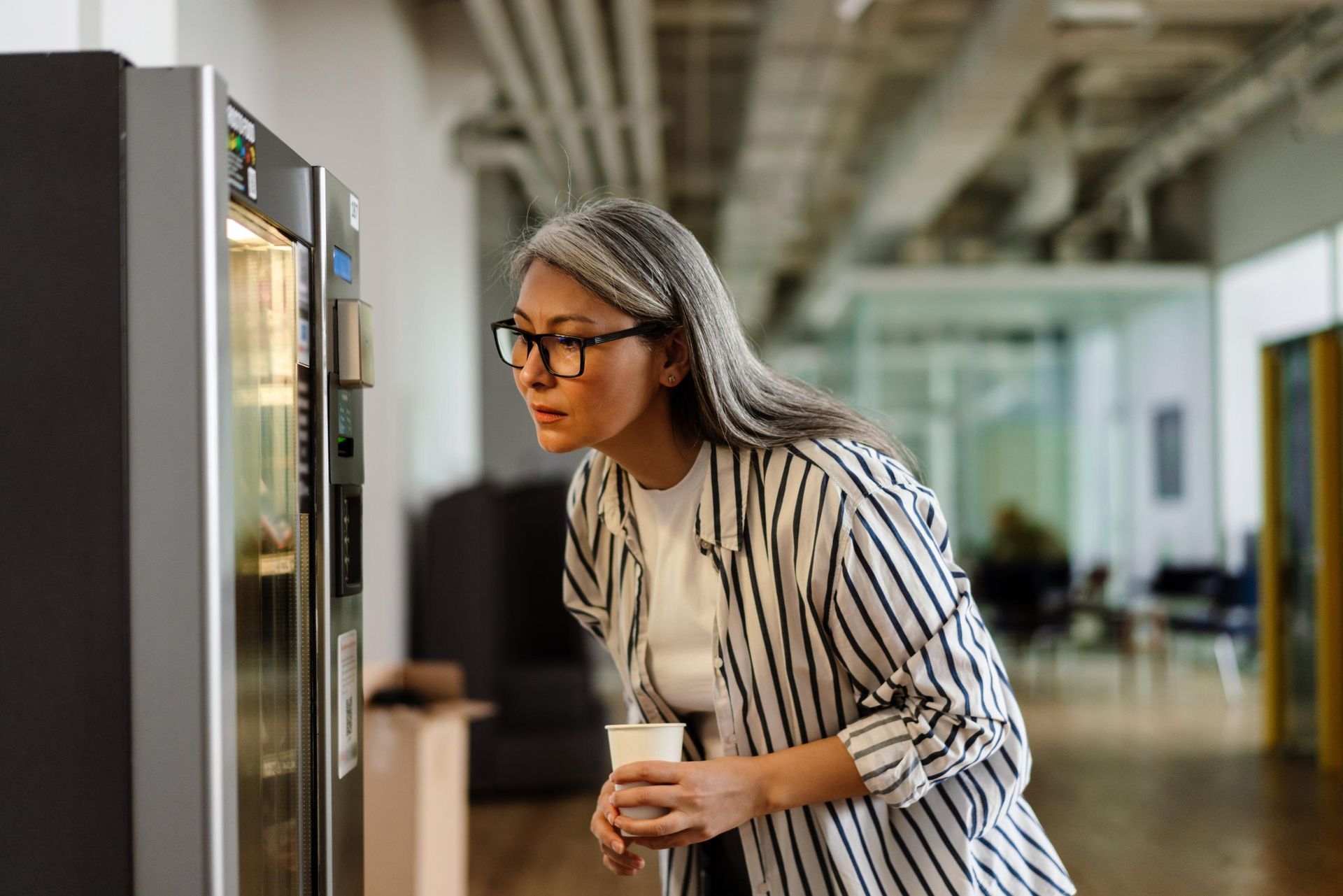 Woman looking at vending machine with a coffee cup in a modern office.