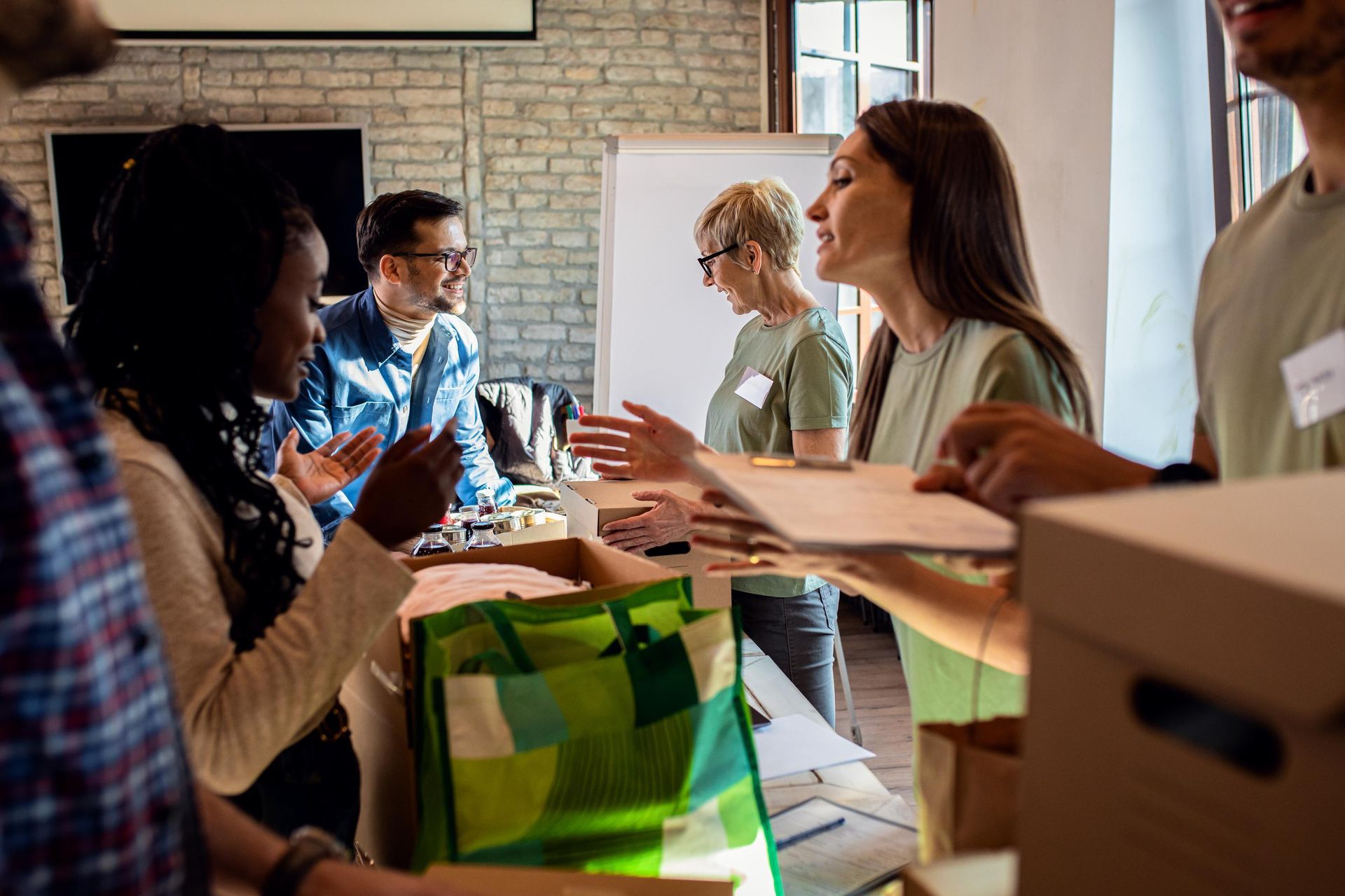 People volunteering, interacting at a table with bags, supplies. Bright room, some wearing volunteer shirts.
