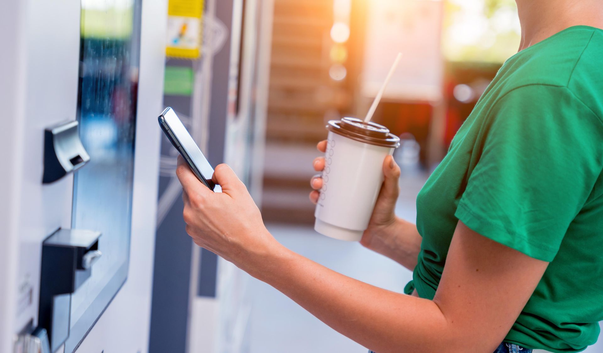 Woman using phone to pay at a vending machine, holding coffee cup. Bright sunlight in the background.