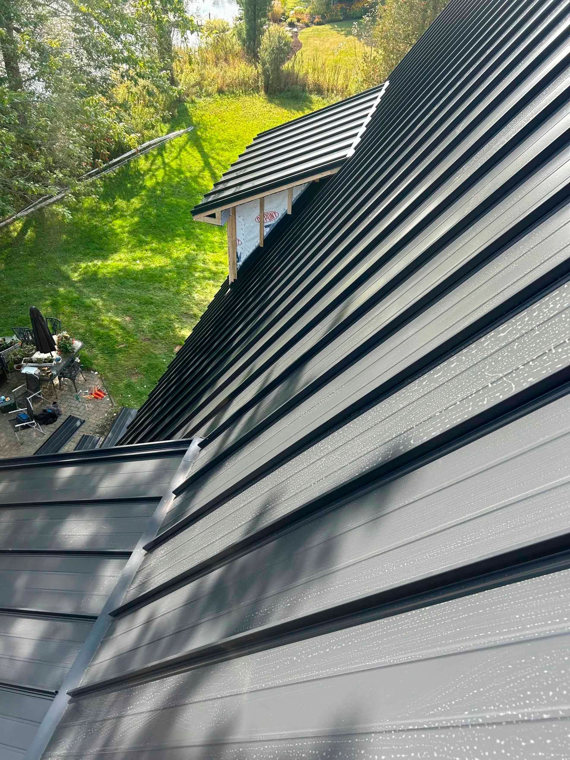 Toit en métal noir percé de gouttelettes d'eau, dans un cadre extérieur ensoleillé.