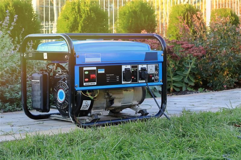 Blue portable generator on a paved surface with a background of green plants and a fence.