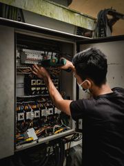 Electrician in mask working on control panel with a drill.