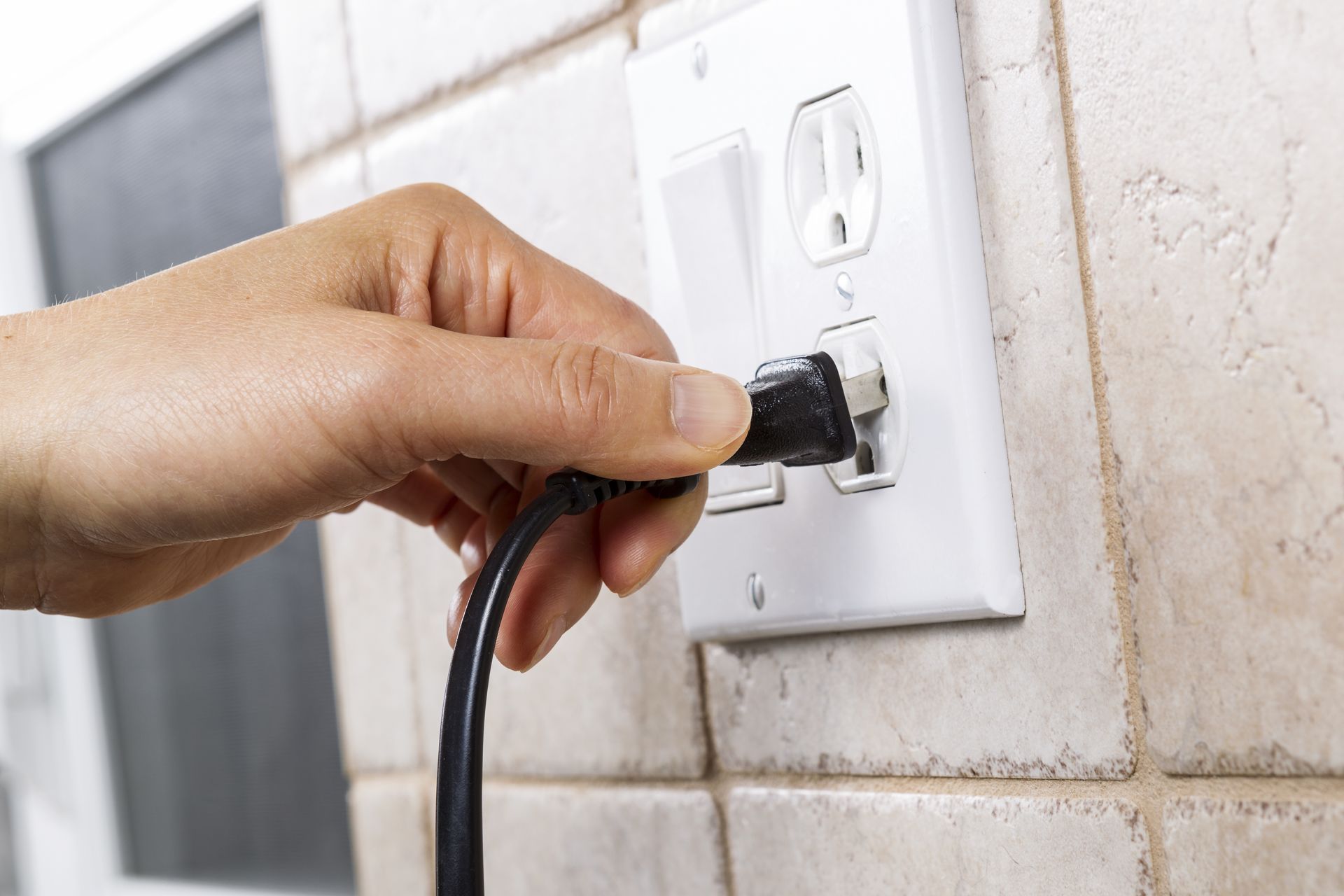 Hand plugging a black power cord into a white electrical outlet on a tiled wall.