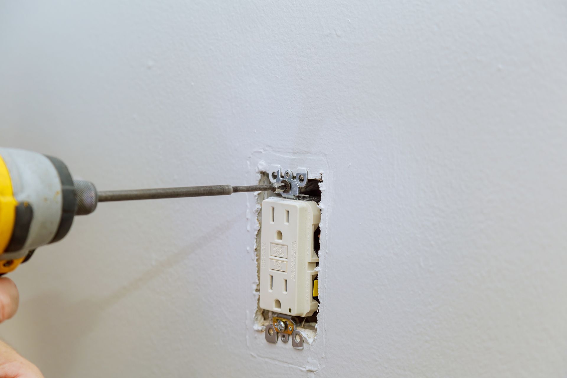 A person is using a power drill to install an electrical outlet in a white wall.