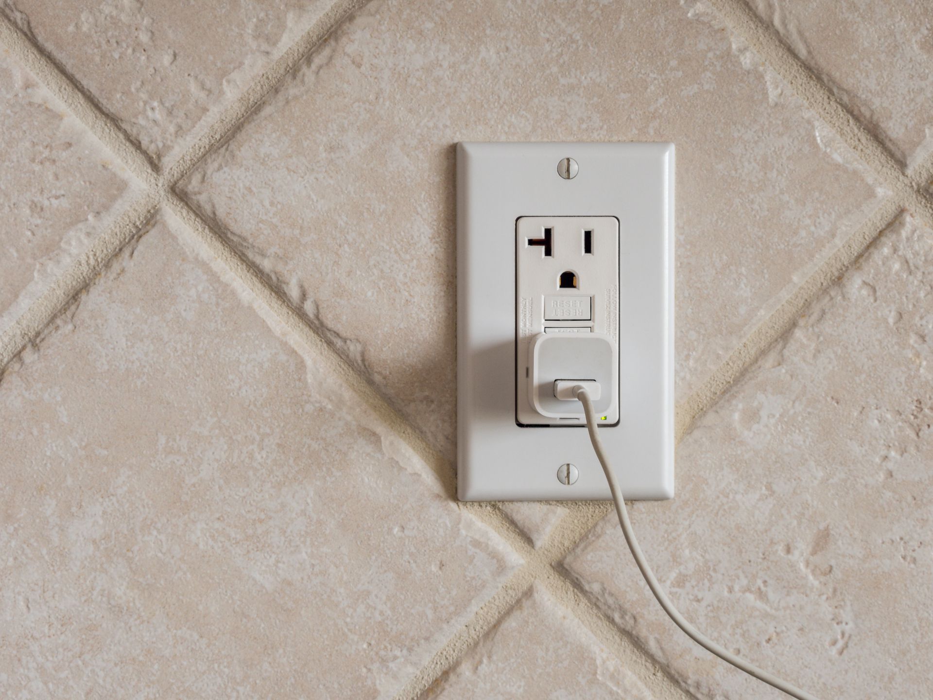 White electrical outlet on beige tile wall, small white charger plugged in.