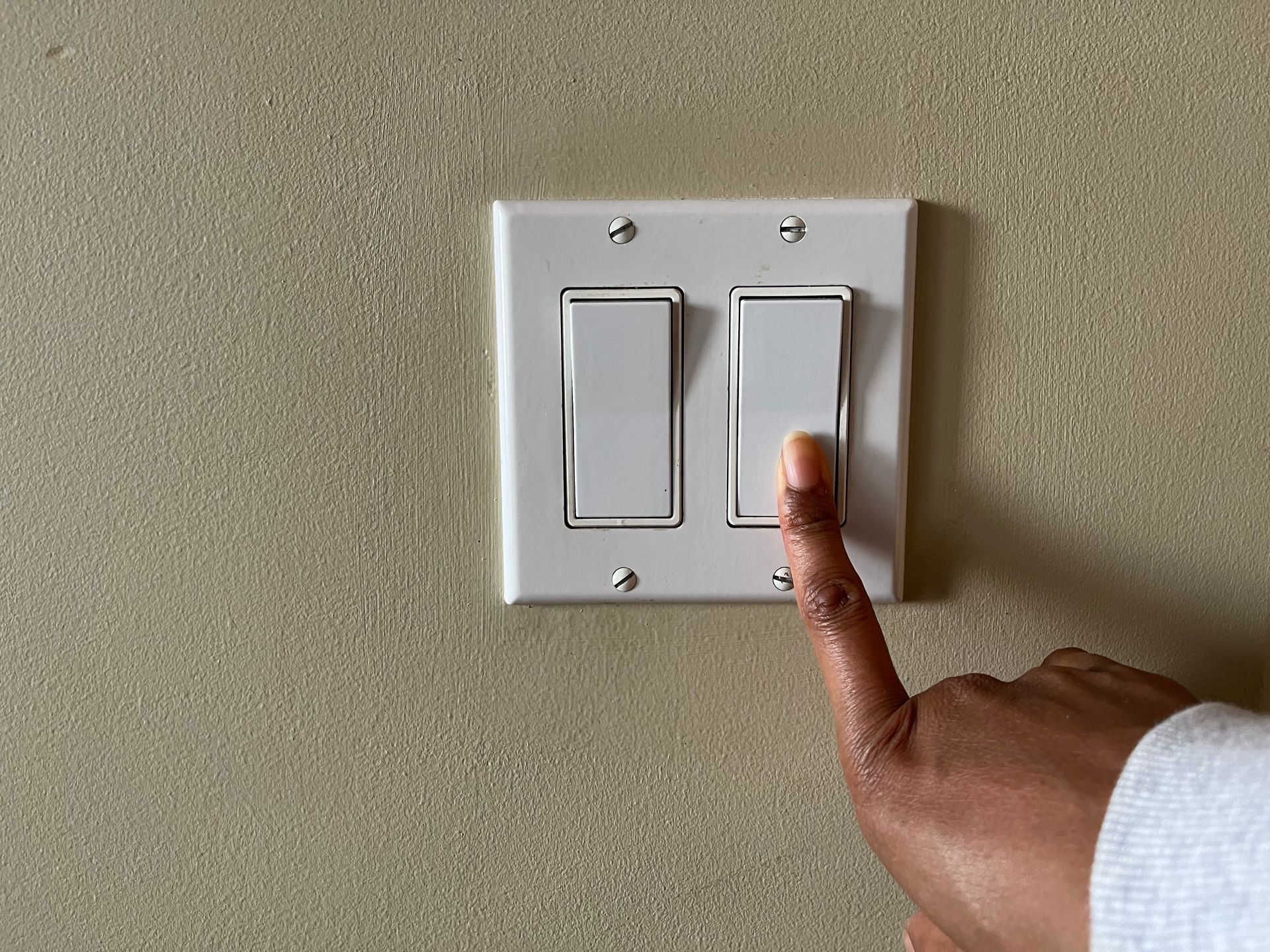 Finger pressing down a light switch. White switch plate on beige wall.