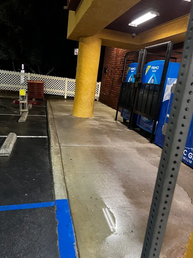 A parking lot at night with a yellow pillar, a white fence, and two blue Primo water vending machines under a lit canopy. Wet pavement and a shopping cart are visible.