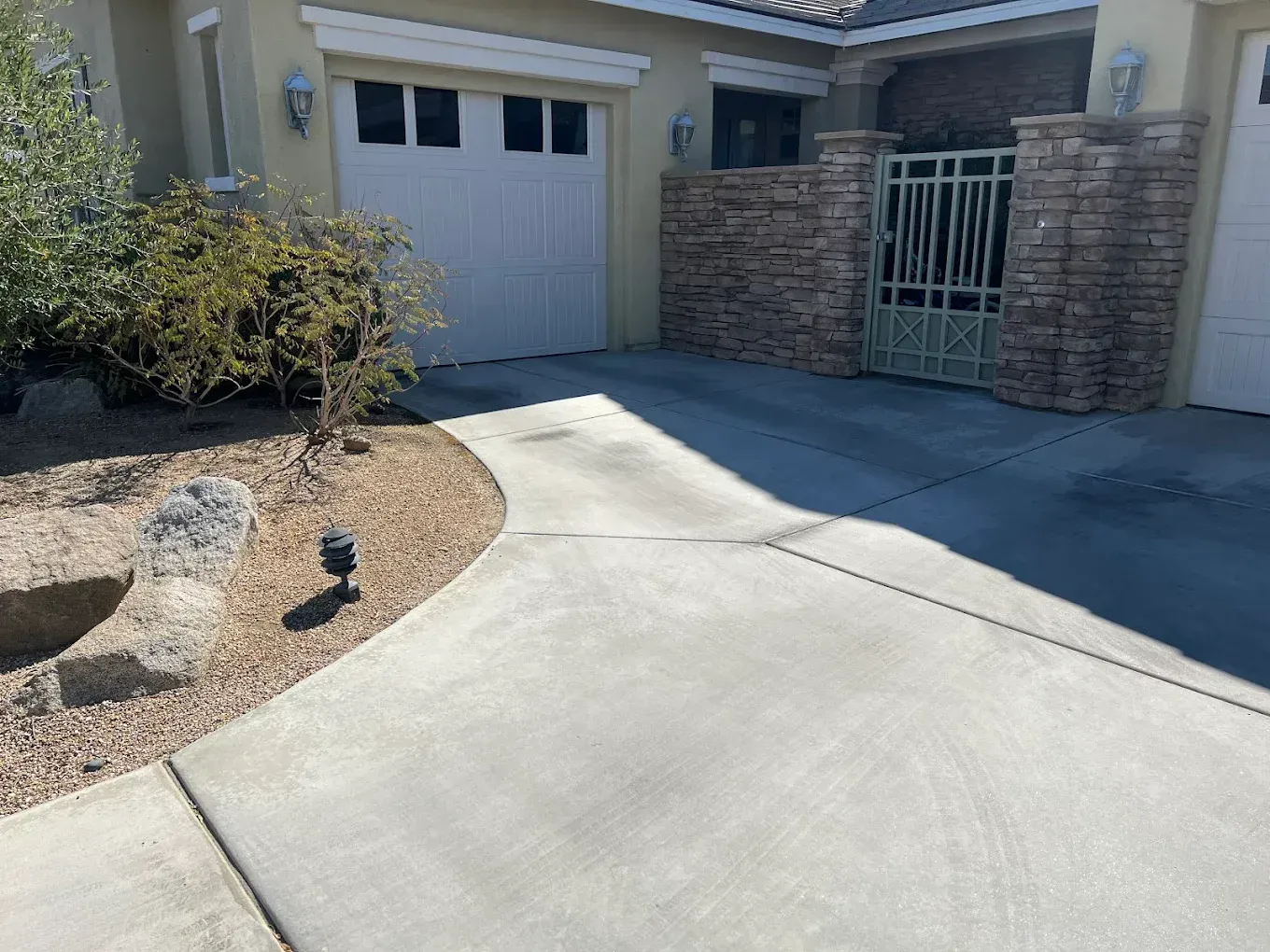 Concrete driveway leading to a two-car garage and gated entryway, with desert landscaping including rocks, gravel, and a small shrub on the left.