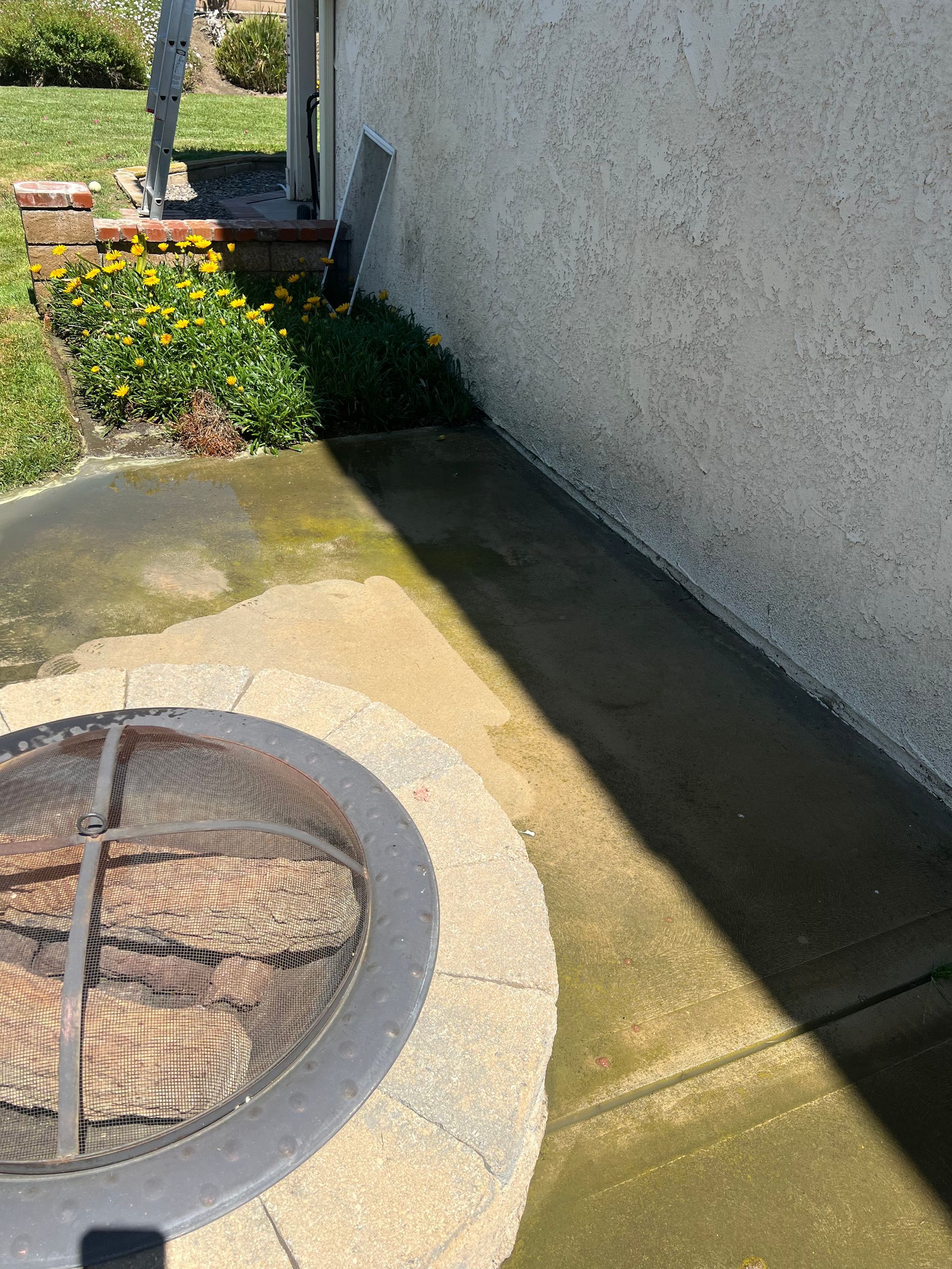 A metal fire pit with a mesh cover sits on a stone base near a stucco wall; yellow flowers and a ladder are in the background, partially shaded.