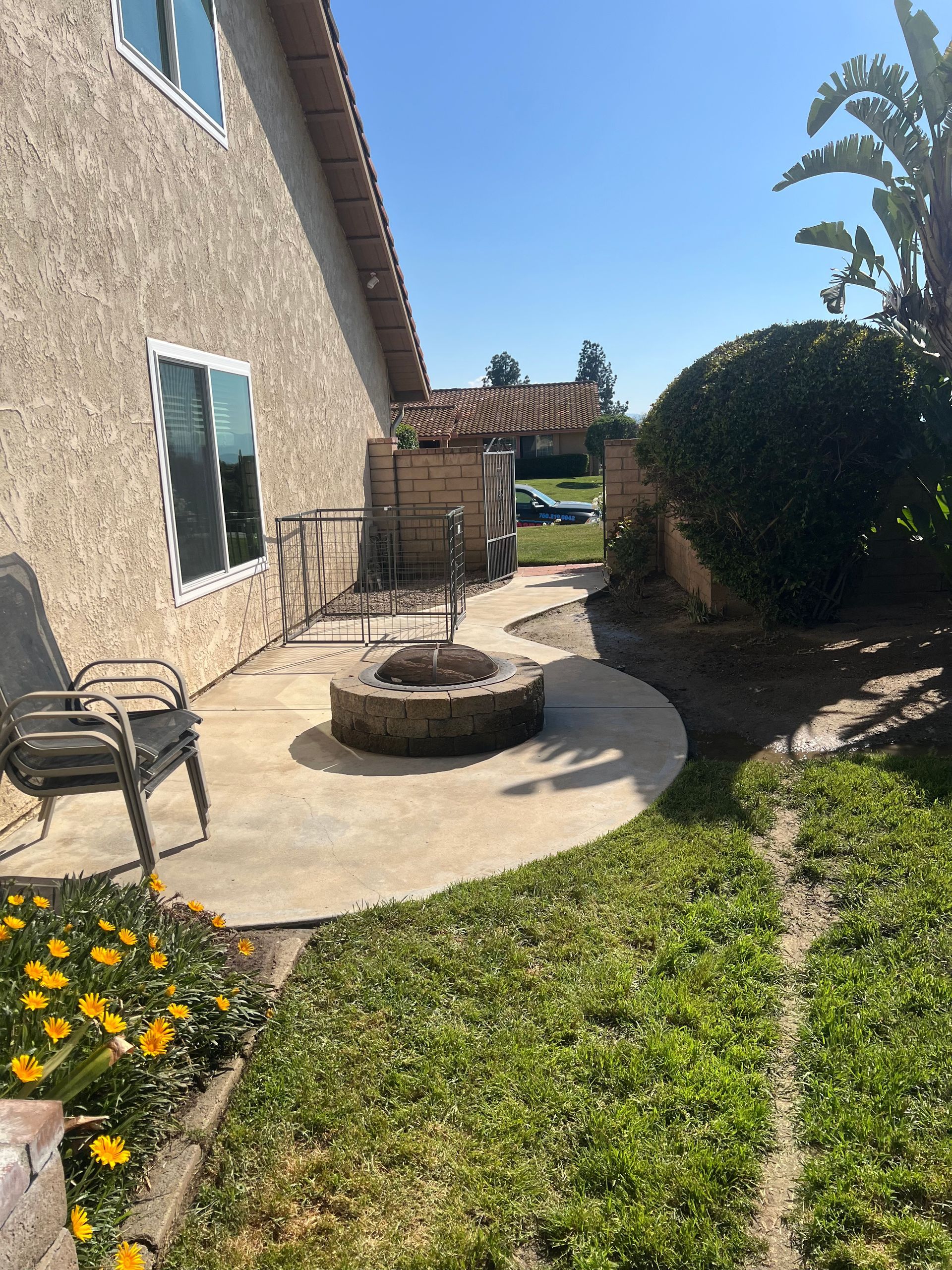 A backyard patio with a fire pit, metal chairs, yellow flowers, and a gated area next to a beige house with a lawn and hedge.