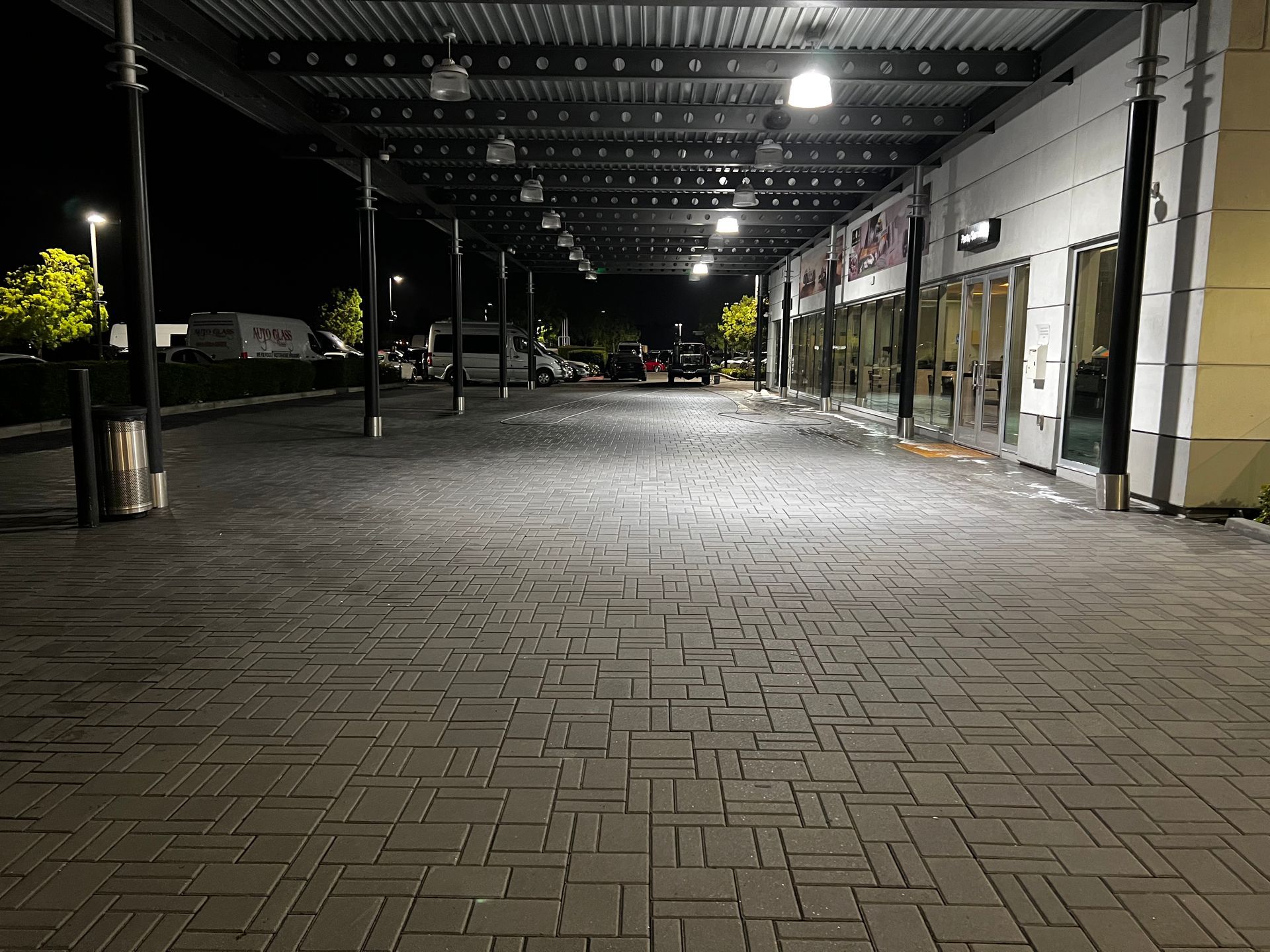 Covered walkway with brick flooring outside a building at night, illuminated by overhead lights, with parked cars and trees visible in the background.