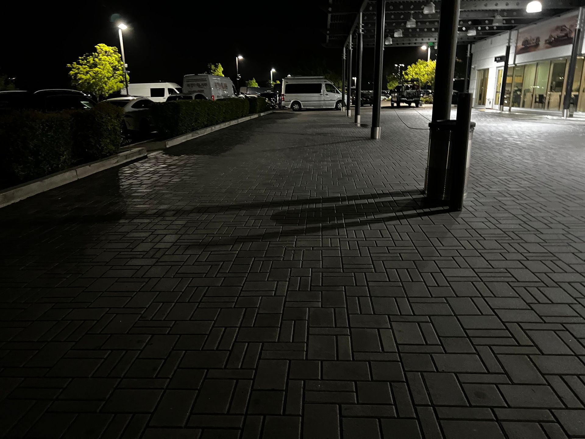 A paved sidewalk at night with cars parked along the side, illuminated by streetlights and the windows of a nearby building.