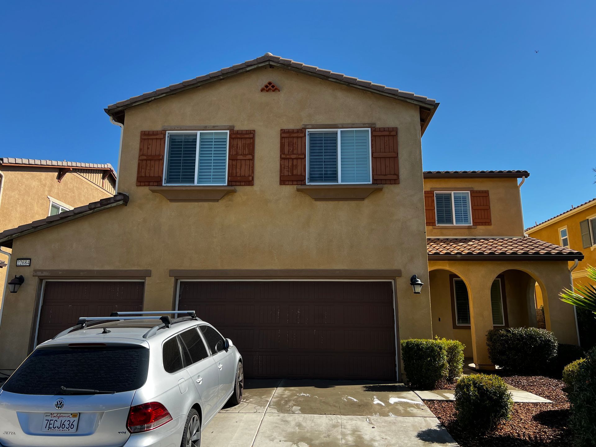 Two-story suburban house with brown garage doors, tan exterior, and a white car parked in the driveway under a clear blue sky.
