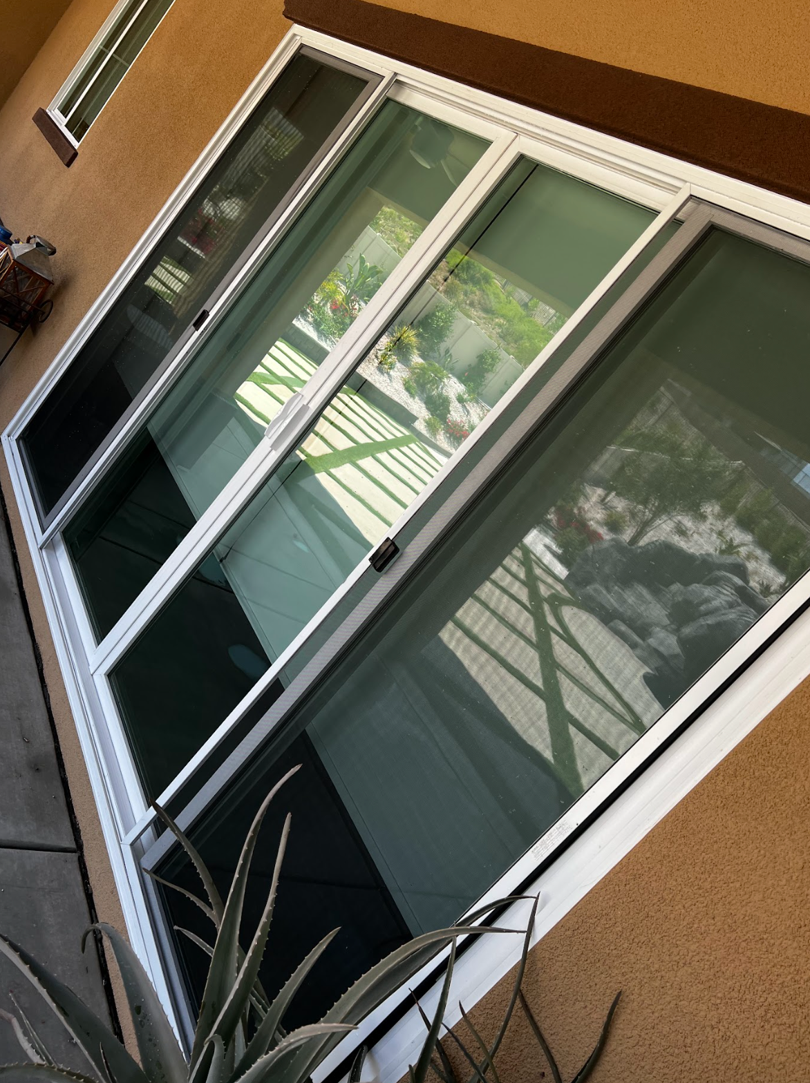 A sliding glass door with closed blinds is set in a tan exterior wall. The door reflects a backyard with a patio cover, garden, and rocks. An aloe plant is visible in the foreground.