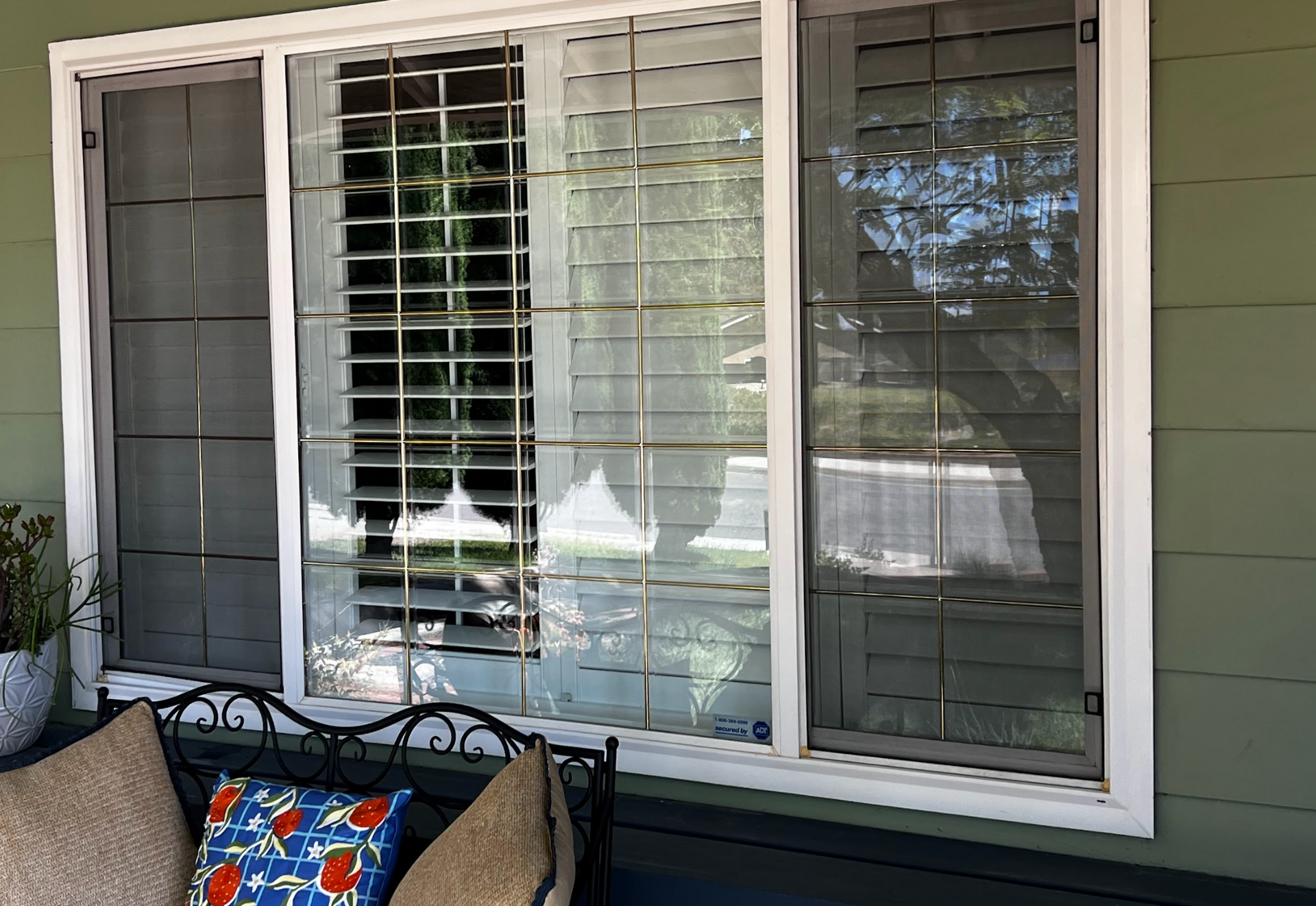 A large white-framed window with closed blinds is behind a black metal bench with patterned cushions on an outdoor porch.