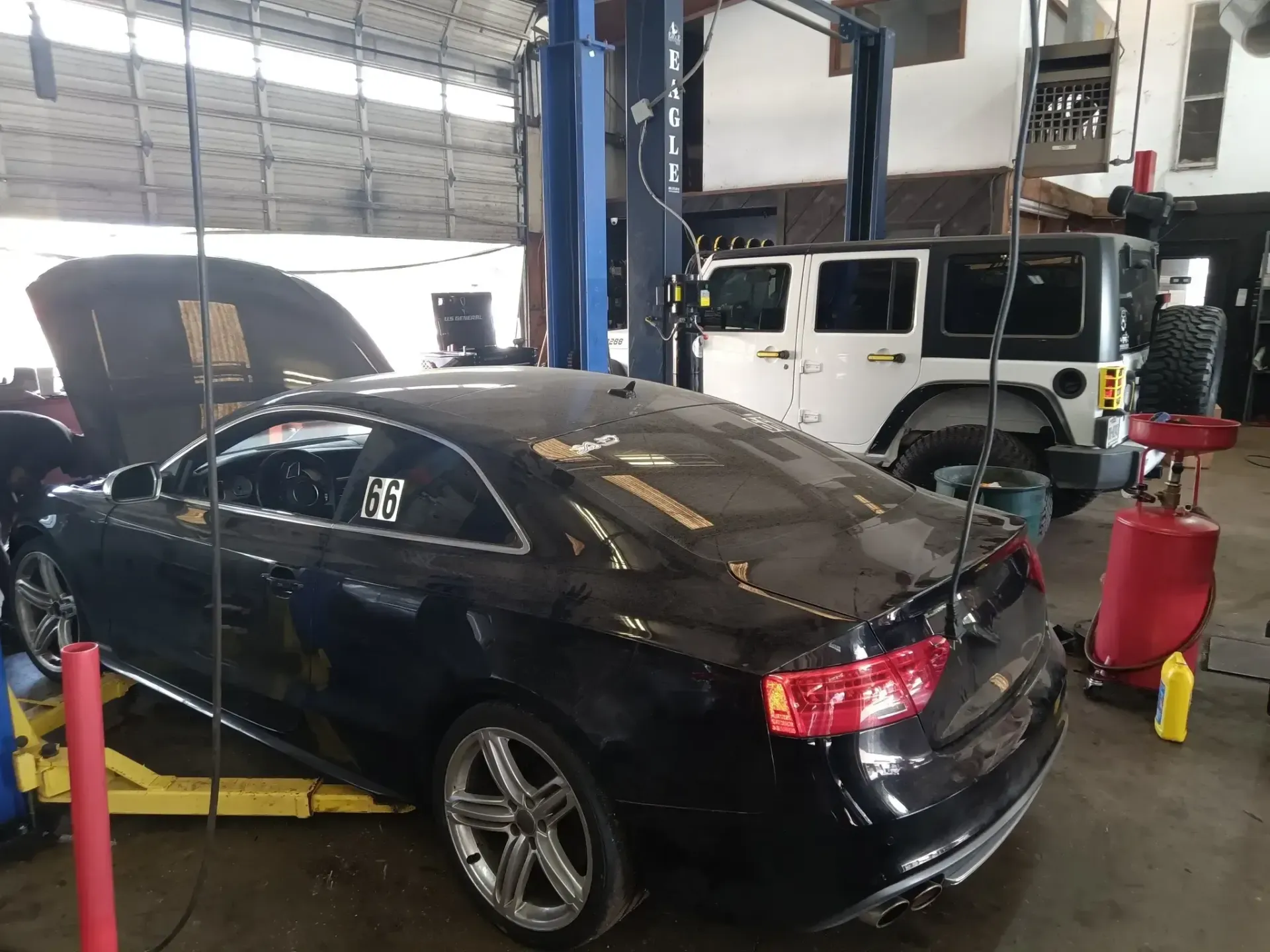 Black car on a lift in a repair shop with a white Jeep in the background. Hood open. | We Fix Cars