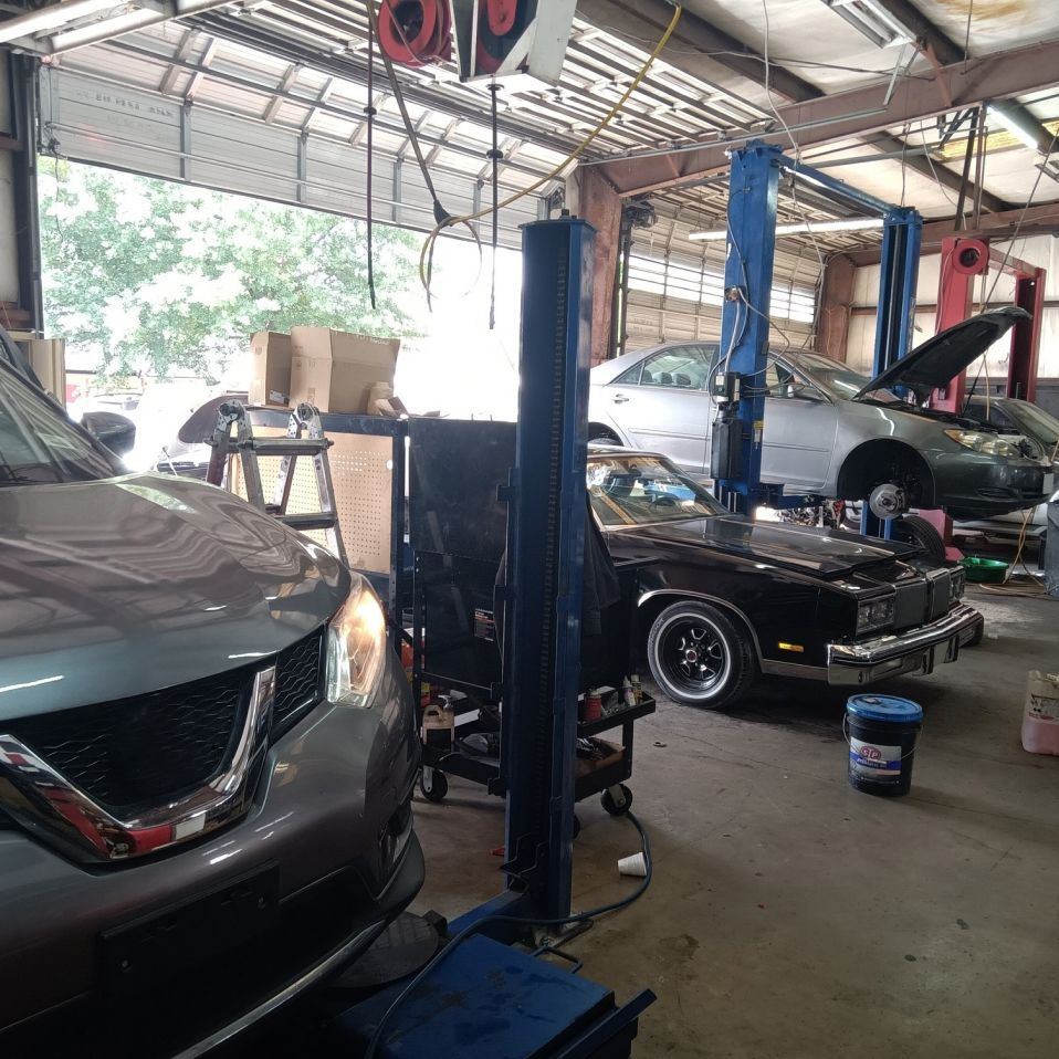 Inside a car repair shop with vehicles on lifts; a gray SUV and a classic black car are in focus. | We Fix Cars