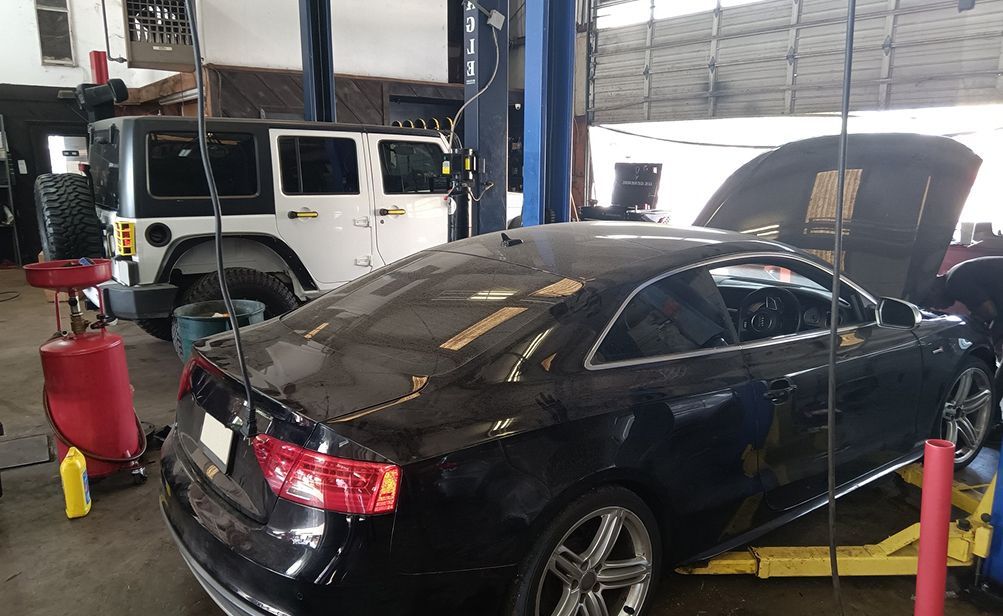Black car raised on a lift in a repair shop, hood open. White Jeep parked nearby. | We Fix Cars