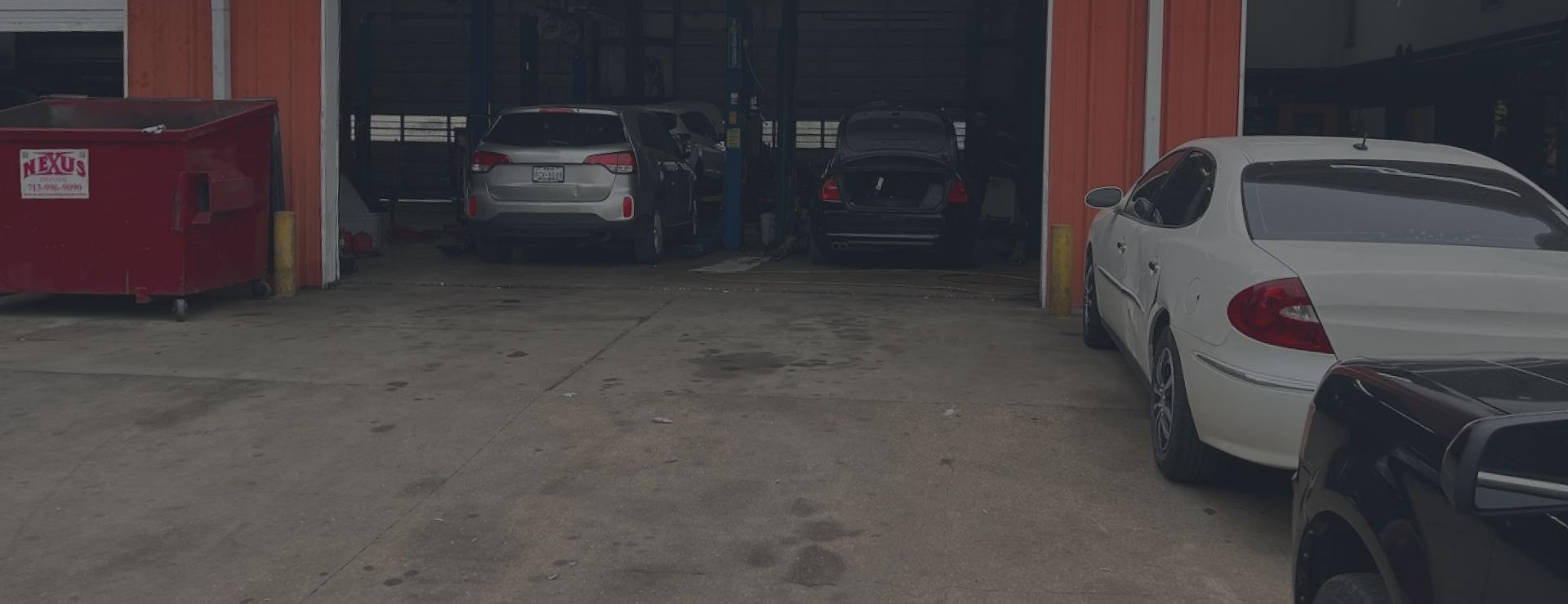 Cars parked in front of an auto repair shop. A red dumpster is visible on the left. | We Fix Cars