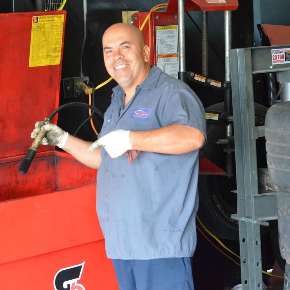 Mechanic in blue uniform pointing while holding tool, next to red machinery. | We Fix Cars