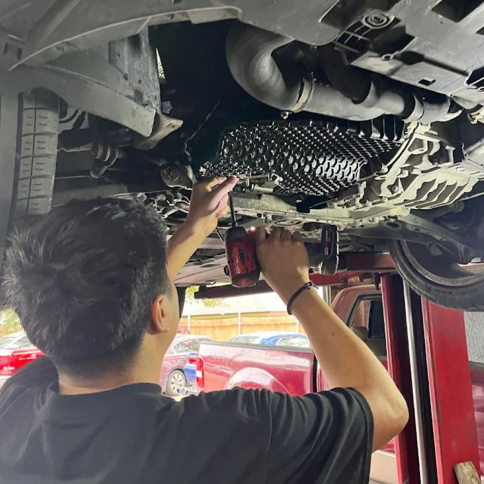 Mechanic working under a car, using a power tool to remove a component. The car is on a lift. | We Fix Cars