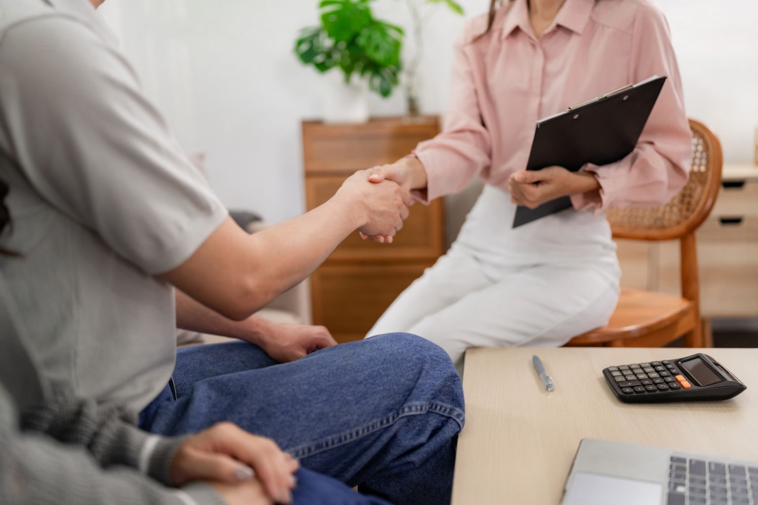 Woman in pink shirt shakes hands with a person in a gray shirt, seated near a calculator, and laptop.