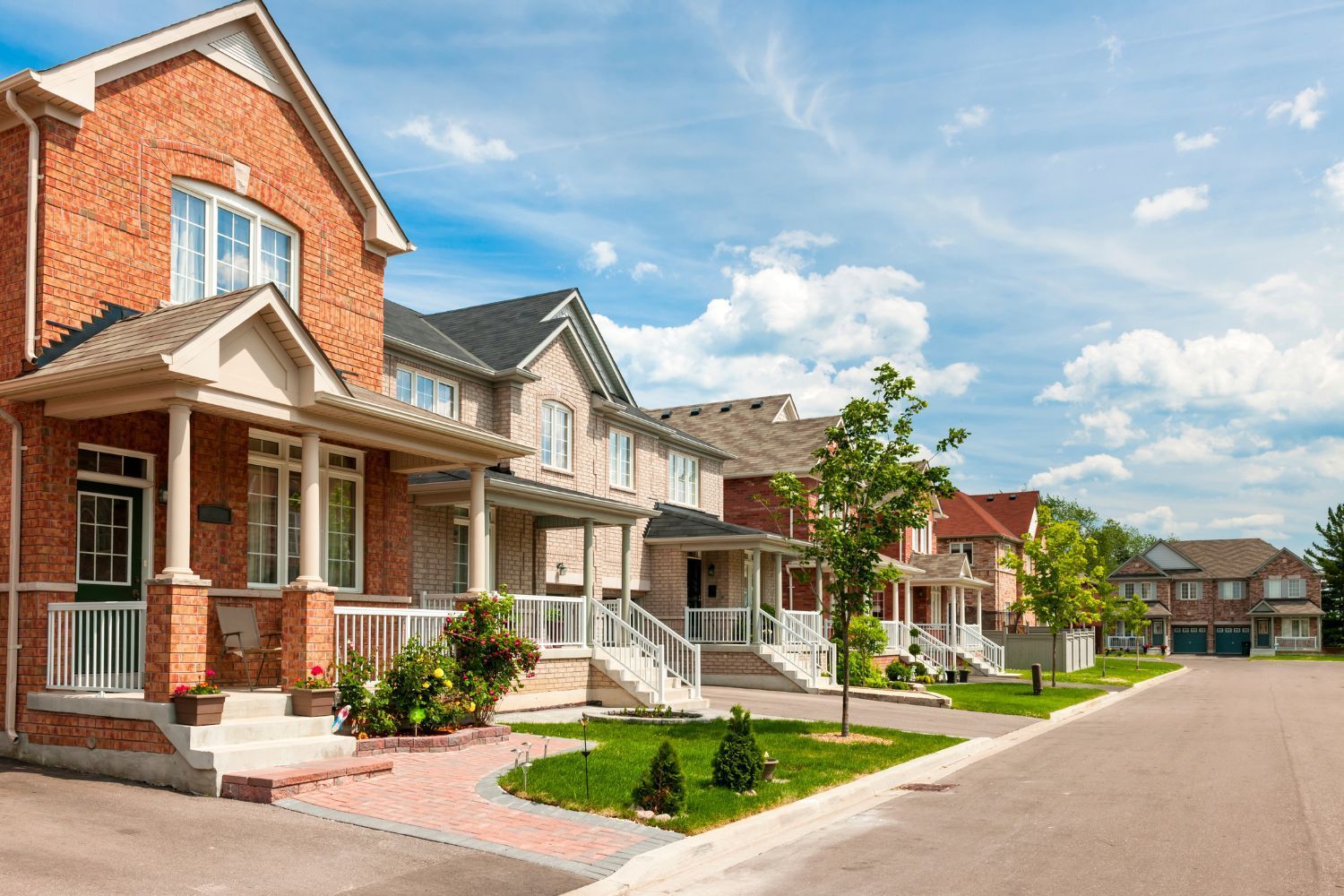 Row of brick and stone houses on a sunny street, with front porches and green lawns.