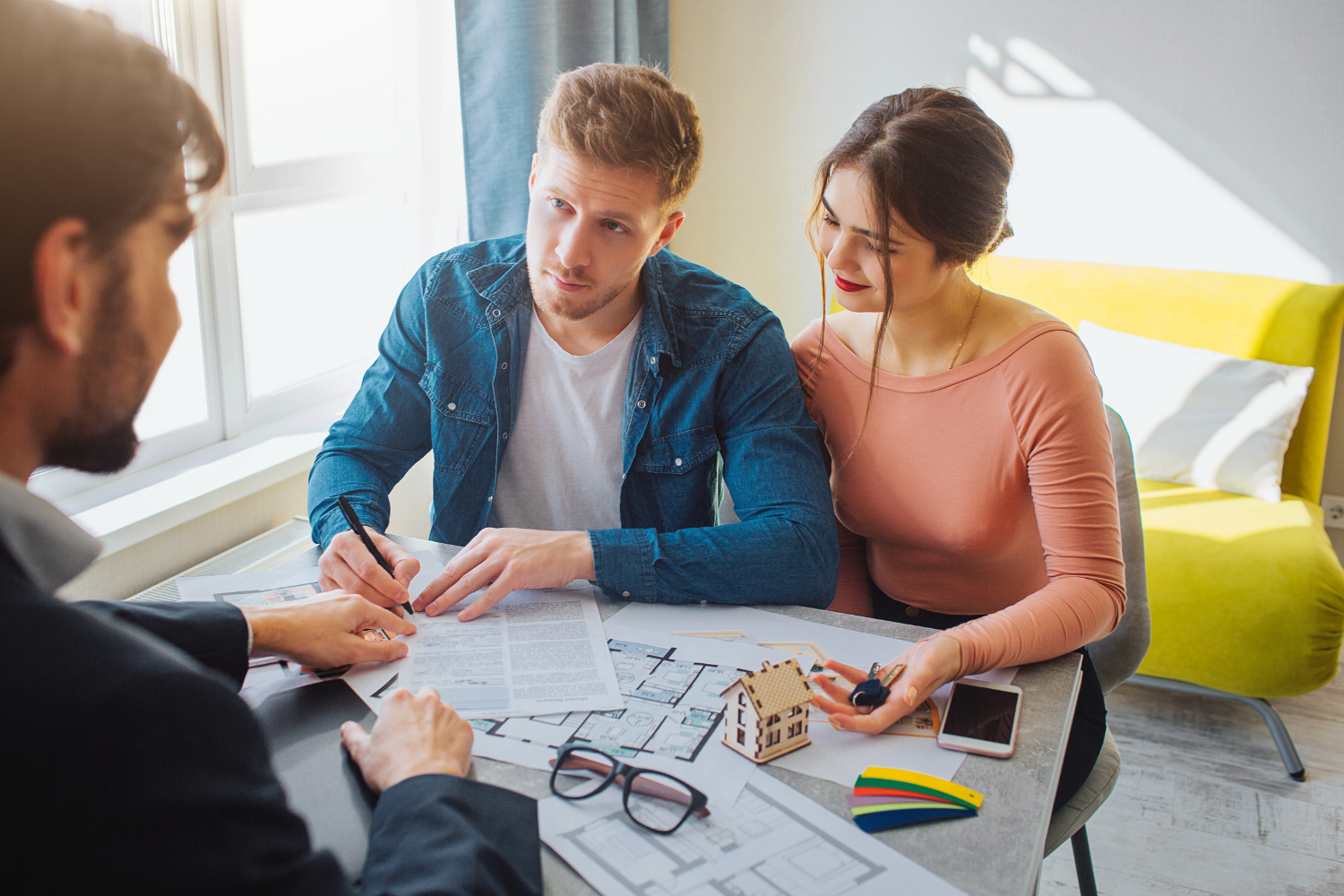 Couple signing papers with a person in a meeting, with house model and keys on table.