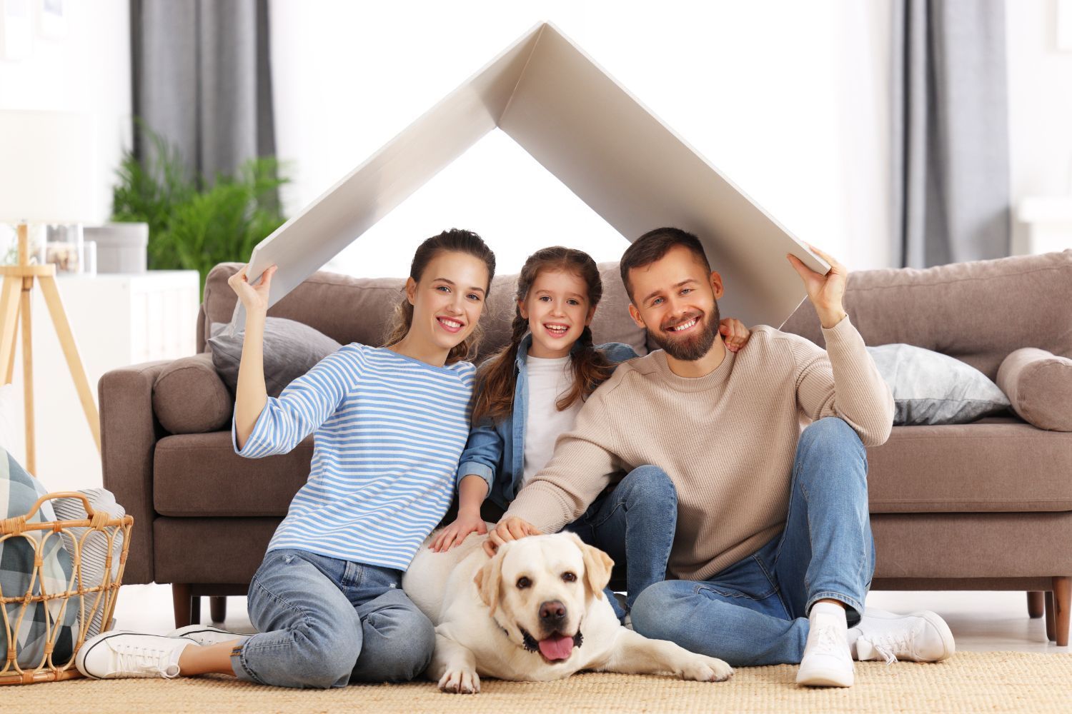 Family of three and dog sit under a cardboard roof in a living room; smiling.